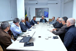 Reitor Paulo César Montagner sentado à cabeceira de mesa de reuniões, tendo aos seus dois lados representantes da Unicamp e do Serviço Nacional de Aprendizagem Industrial (Senai), e, ao fundo, um quadro com uma fotografia aérea noturna do campus da Universidade em Campinas.