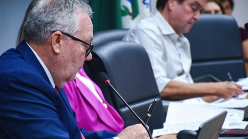 Reitor Paulo Cesar Montagner fala ao microfone sentado à mesa diretora da sala do Conselho Universitário (Consu), tendo ao seu lado esquerdo dois pró-reitores, uma mulher e um homem, que integram a sua equipe de gestão.