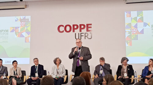 Reitor Paulo César Montagner discursa de pé em palco de auditório na Universidade Federal do Rio de Janeiro (UFRJ). Atrás dele, também no palco, estão sentados de frente para a plateia outros sete participantes do Fórum de Reitores do Brics+.