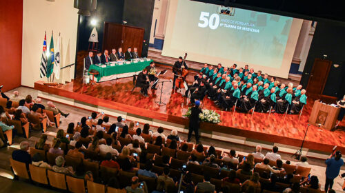 Vista do maior auditório da Faculdade de Ciências Médicas (FCM) da Unicamp a partir do alto da plateia, que está lotada. No palco, há, à esquerda, a mesa de autoridades, à qual estão sentadas sete pessoas; no centro, um quarteto de músicos e cinco fileiras de cadeiras, todas ocupadas por antigos alunos da oitava turma do curso de medicina; e, à direita, o púlpito de onde fala a mestre de cerimônias.