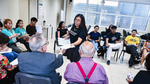 O reitor da Unicamp, Paulo Cesar Montagner, o coordenador geral da Universidade, Fernando Coelho, e mais de dez estudantes indígenas estão sentados em cadeiras posicionadas em círculo em uma sala de aula. O reitor recebe um documento das mãos de uma estudante que está de pé no centro da roda.