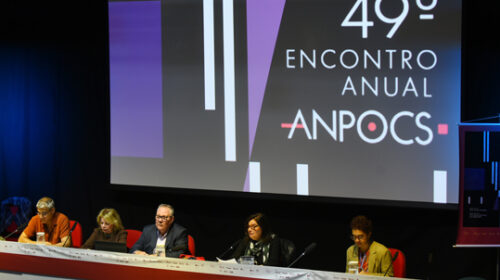 Vista diagonal da mesa de autoridades de um auditório, à qual estão sentadas, em cadeiras vermelhas, três mulheres e dois homens. O reitor da Unicamp, Paulo Cesar Montagner, está na posição central. Ao fundo, um telão exibe o nome do evento: 49º Encontro Anual Anpocs.