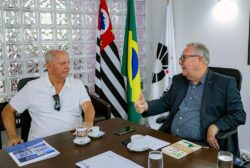 O reitor da Unicamp, Paulo Cesar Montagner, à direita, e o presidente da Confederação Nacional dos Clubes, Arialdo Boscolo, conversam sentados a uma mesa de reuniões. O reitor ocupa o assento da cabeceira. Atrás dele há uma panóplia com as bandeiras Estado de São Paulo, do Brasil e da Unicamp.