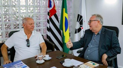 O reitor da Unicamp, Paulo Cesar Montagner, à direita, e o presidente da Confederação Nacional dos Clubes, Arialdo Boscolo, conversam sentados a uma mesa de reuniões. O reitor ocupa o assento da cabeceira. Atrás dele há uma panóplia com as bandeiras Estado de São Paulo, do Brasil e da Unicamp.
