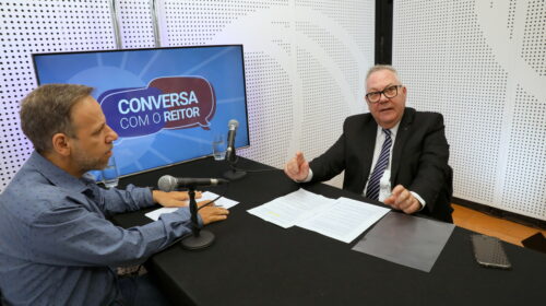 O reitor da Unicamp, Paulo Cesar Montagner, à direita da imagem, e um jornalista, à esquerda, estão sentados a uma mesa, um de frente para o outro, em um estúdio de televisão. A mesa está encostada em uma parede, na qual há um monitor que exibe o logotipo do programa que está sendo gravado, chamado “Conversa com o Reitor”.